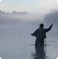 A lone fisherman, knee deep in water casts his line into the foggy waters.