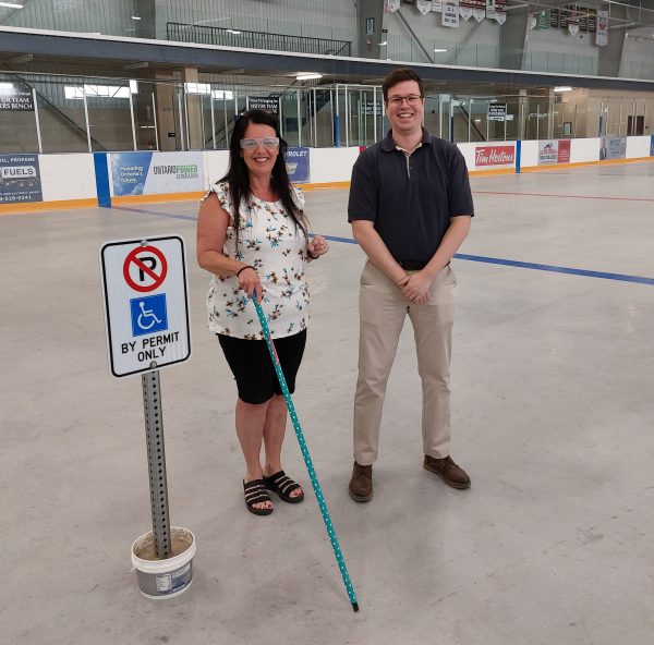 A woman wearing low visibility glasses and a walking stick poses next to a man and an accessible parking sign. 